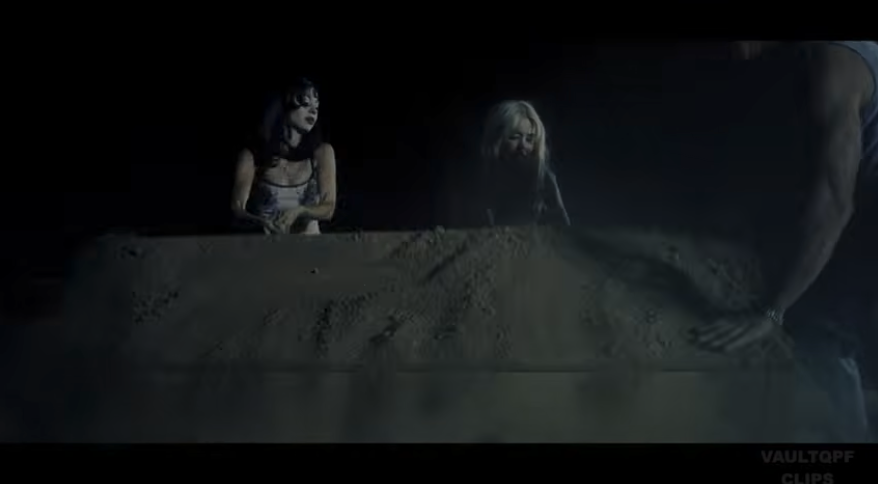 Wide shot: a coffin breaks the surface of the grave at night; two figures at the rim.